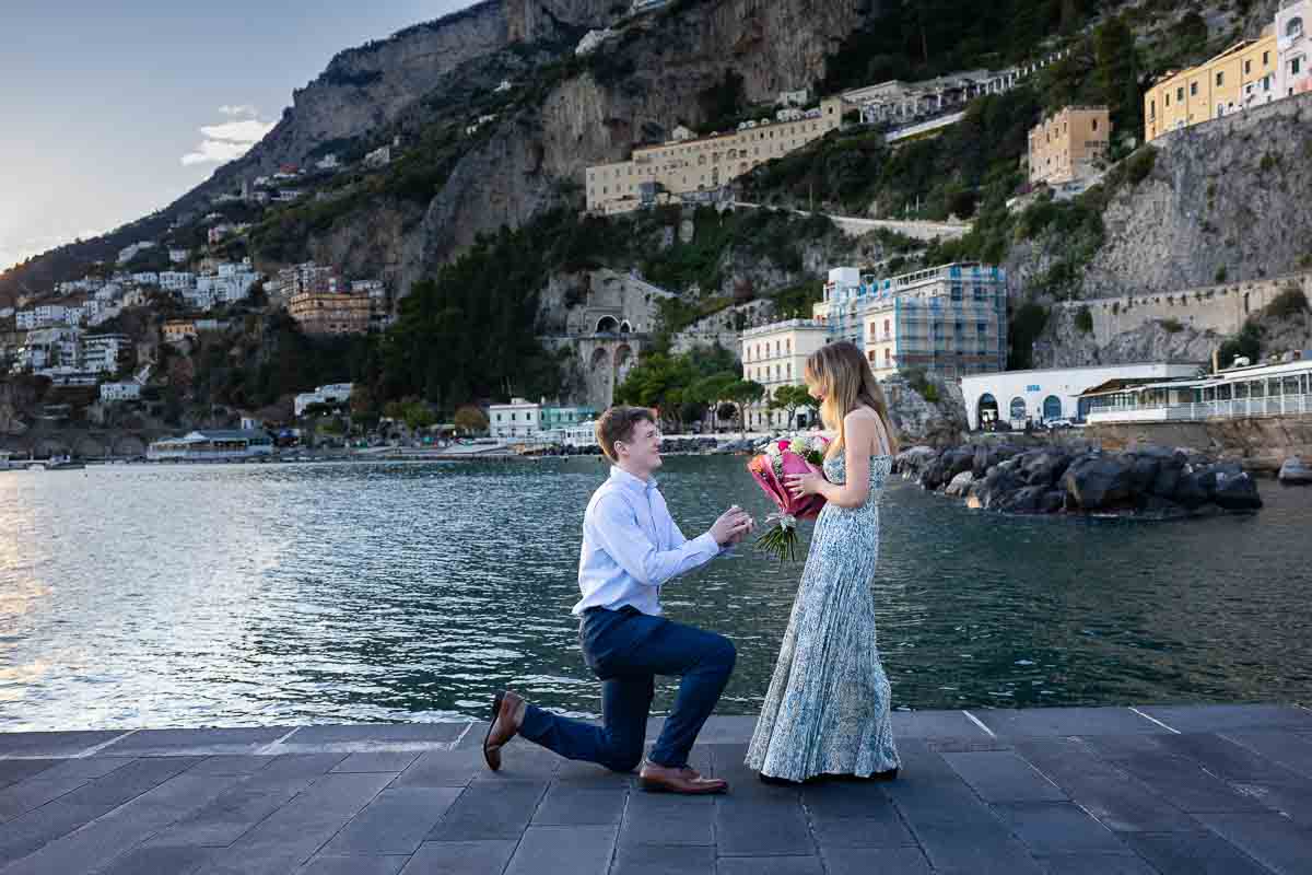 The knee down moment of the wedding proposal photographed in the town of Amalfi coast pier next to water 