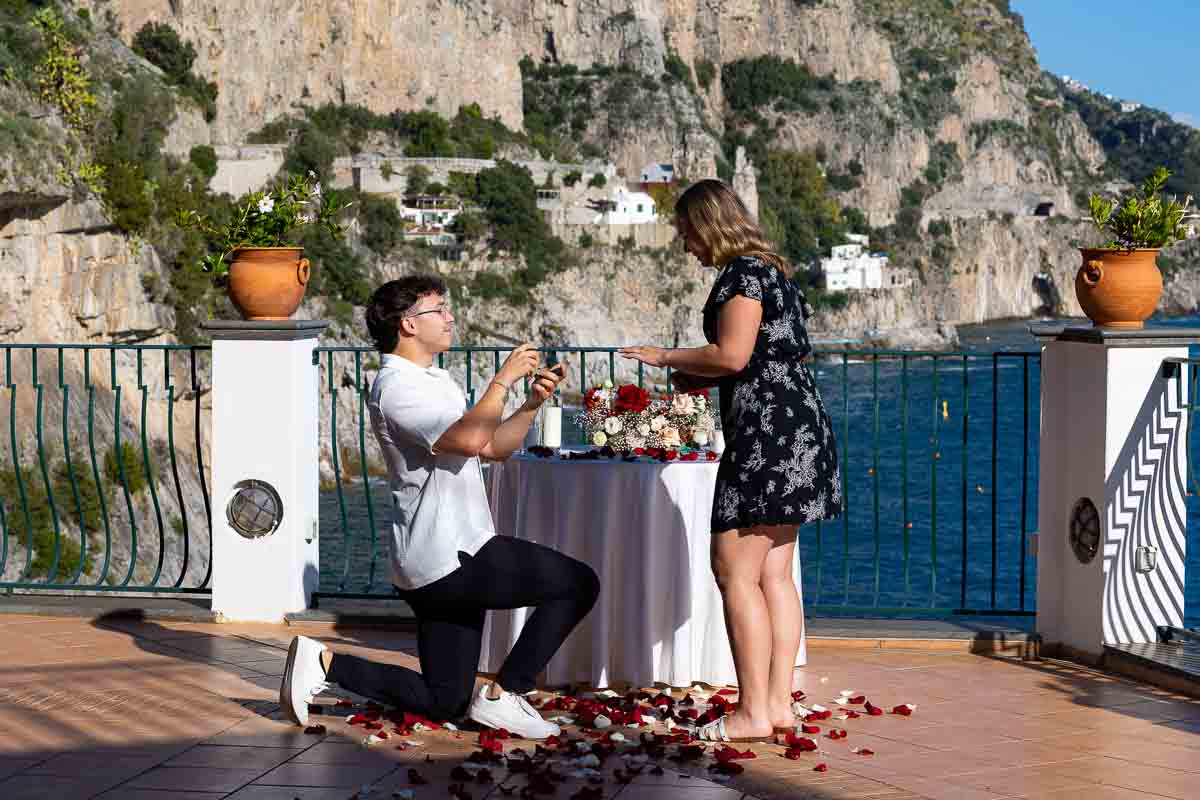 Kneeling down for surprise proposal photography on a private hotel terrace with flowers and beautiful coastal views 