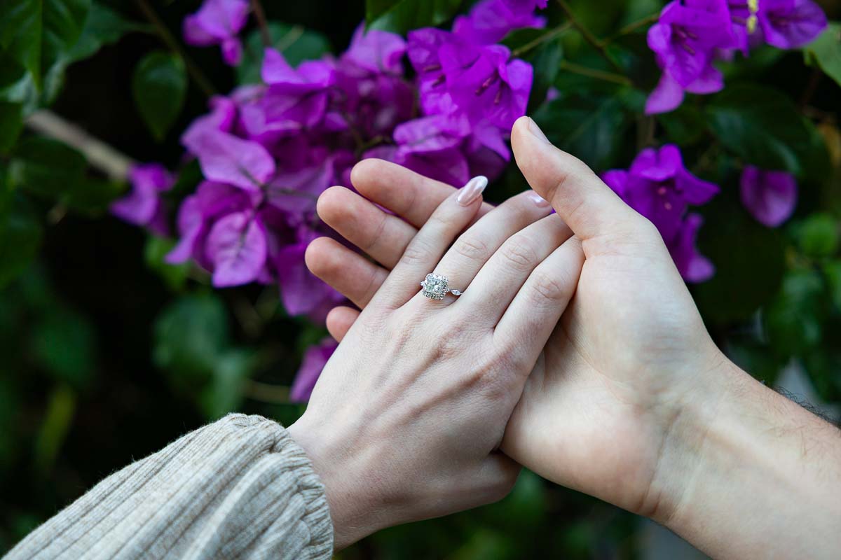 The engagement ring on her hand while holding his over fuchsia flowers