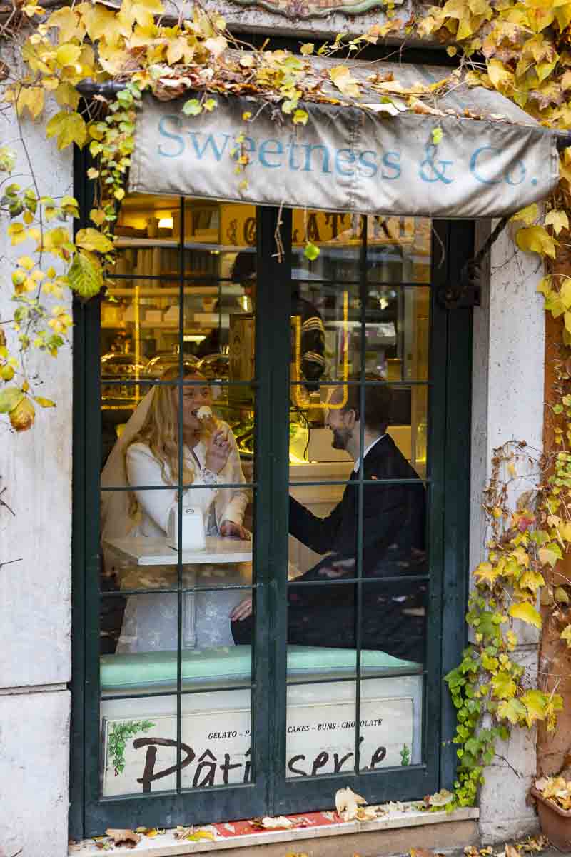 Groom and bride eating ice cream inside a parlor with photographer taking a picture from the outside window pane 
