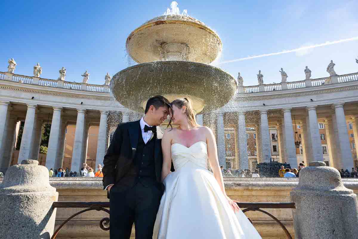 Newlyweds posing forehead to forehead by a classic water fountain in Saint Peter’s Square.