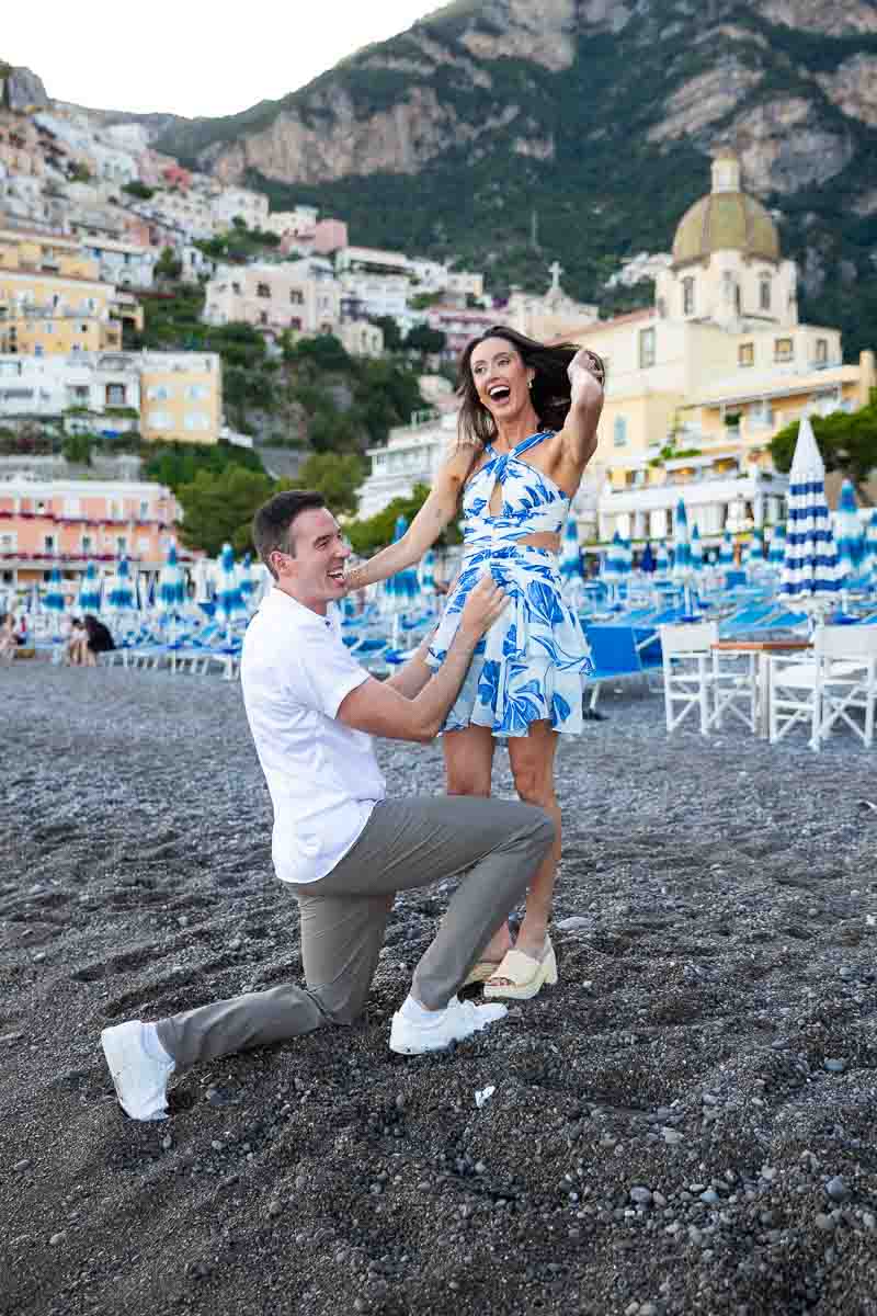 surprised by a wedding marriage proposal asked on the beach of positano on marina grande with the town expanding vertically behind them 