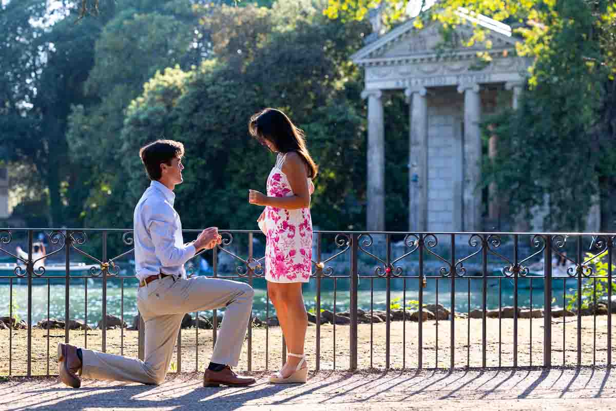 Man kneeling on one knee and proposing as a surprise engagement in front of a roman temple in the park of Villa Borghese by the lake 