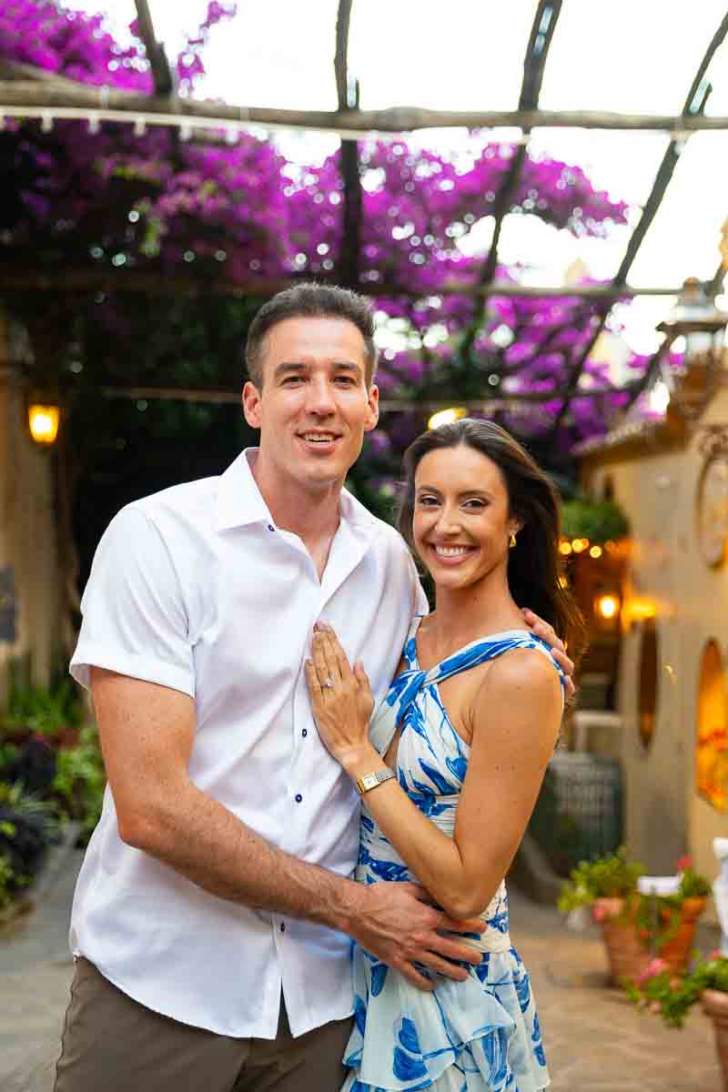 Posed together under a bougainvillea fuchsia plant overhead and smiling looking back at the camera 