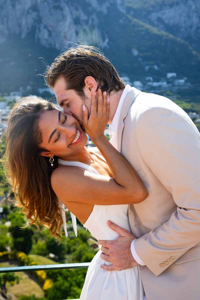 Couple image smiling and being kissed on the cheek during a photographer session using a tele photo lens to capture the candid moment 