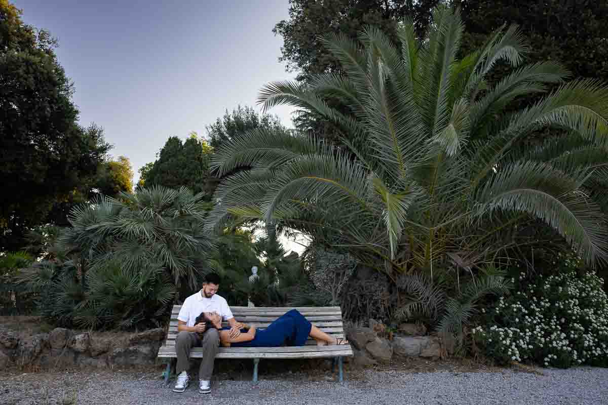 Relaxed couple sitting together on a wooden bench inside Villa Borghese park, captured through a natural lifestyle photoshoot with a peaceful atmosphere surrounded by greenery and soft outdoor light.