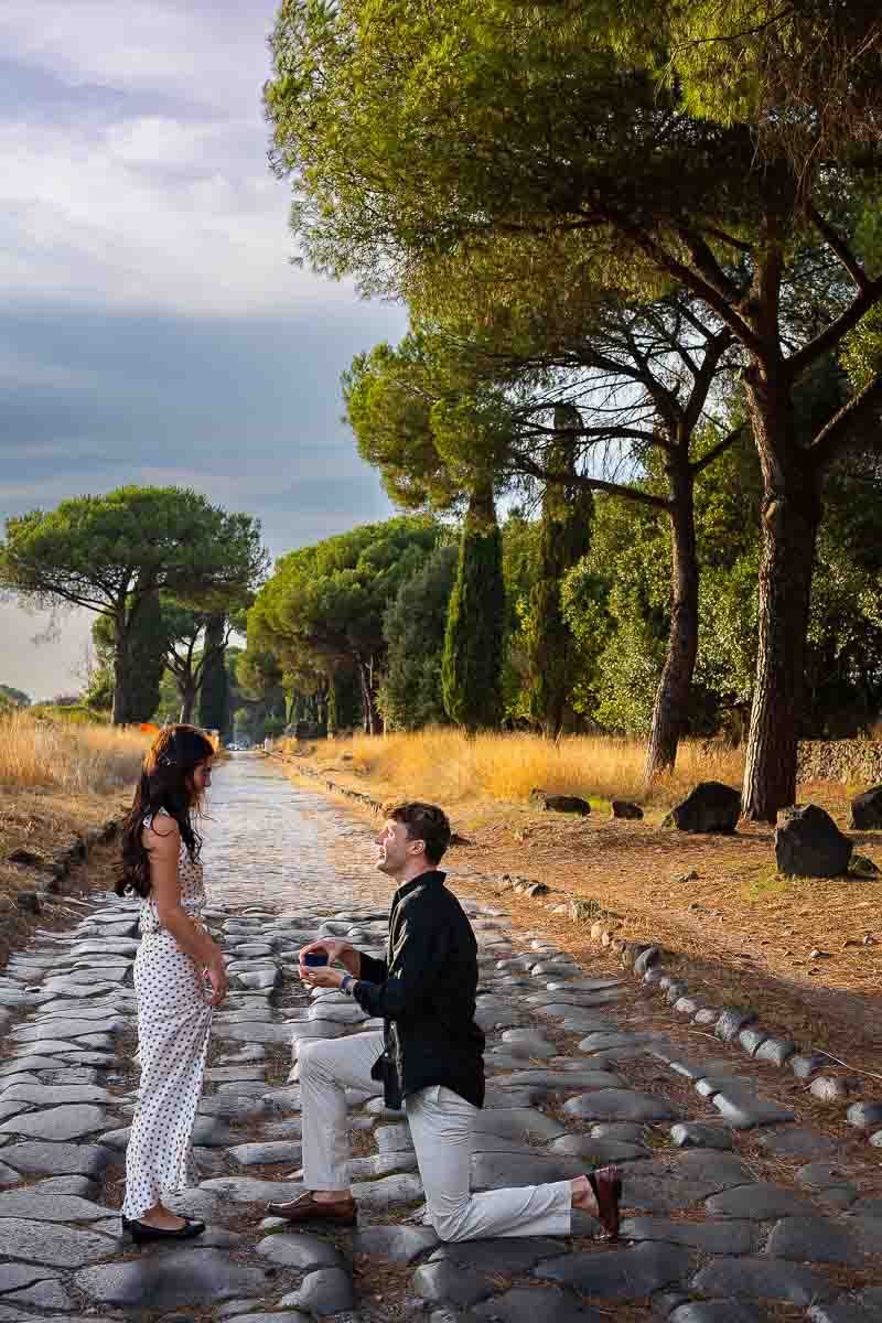 Surprise marriage proposal captured candidly as he kneels before her on the historic roadway, photographed by Andrea Matone Photography in Rome