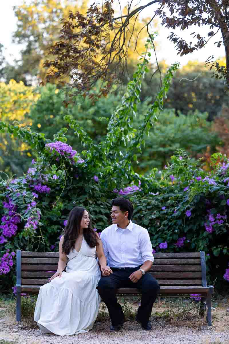 Sitting down on a wooden park bench next to each other looking and taking pictures with a hidden photographer