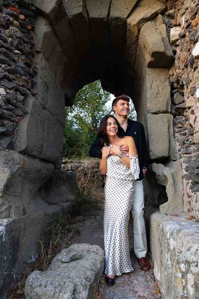 Elegant couple portrait beneath an imposing arched archaeological structure, combining timeless architecture with natural romantic photography