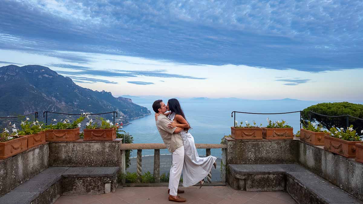 A romantic couple posing for a twilight engagement session overlooking the panoramic Amalfi Coast from a scenic viewpoint inside the historic estate grounds.