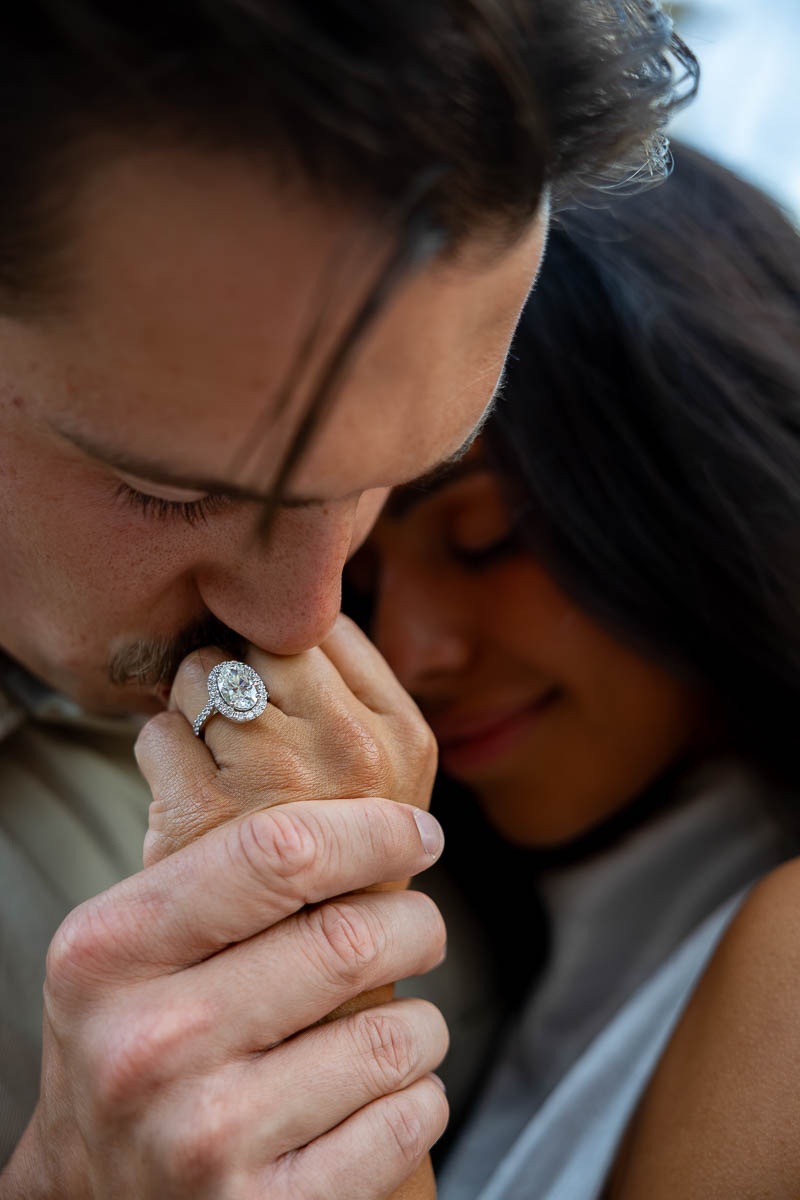 A professional close up photograph showcasing a diamond engagement ring with a soft-focus background of a couple kissing.