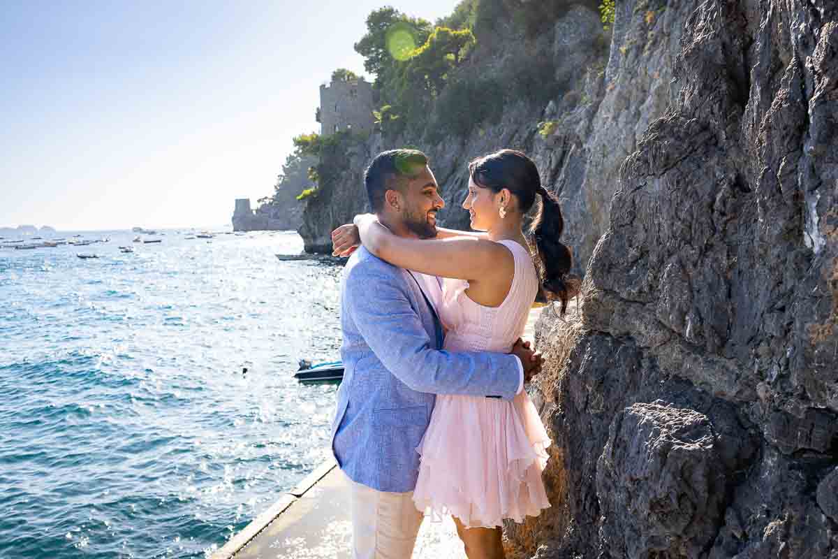Close-up profiles of a couple facing each other beside a rocky cliffside and crashing waves.