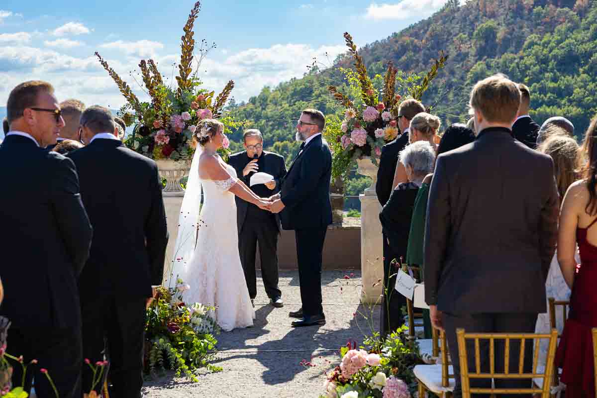 An open-air marriage ceremony taking place high above the rolling Italian countryside.
