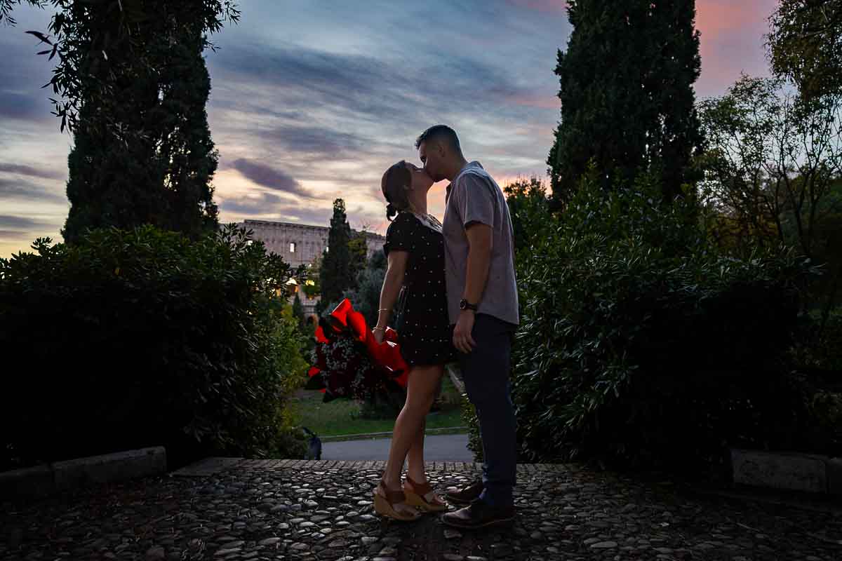 Joyful newlyweds sharing a kiss while holding a beautiful bouquet of crimson roses in the soft twilight light.