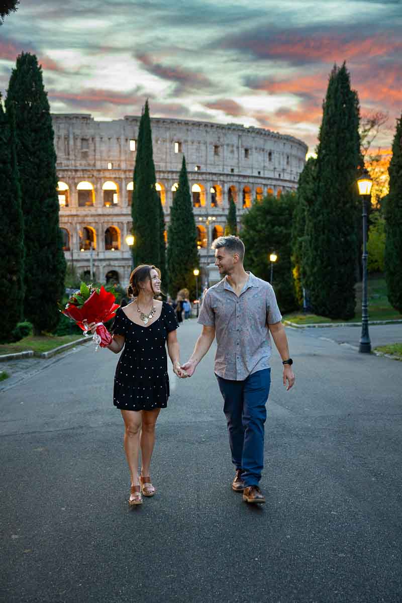 The happy couple walking along a historic Rome walkway with the ancient amphitheater silhouette in the background.