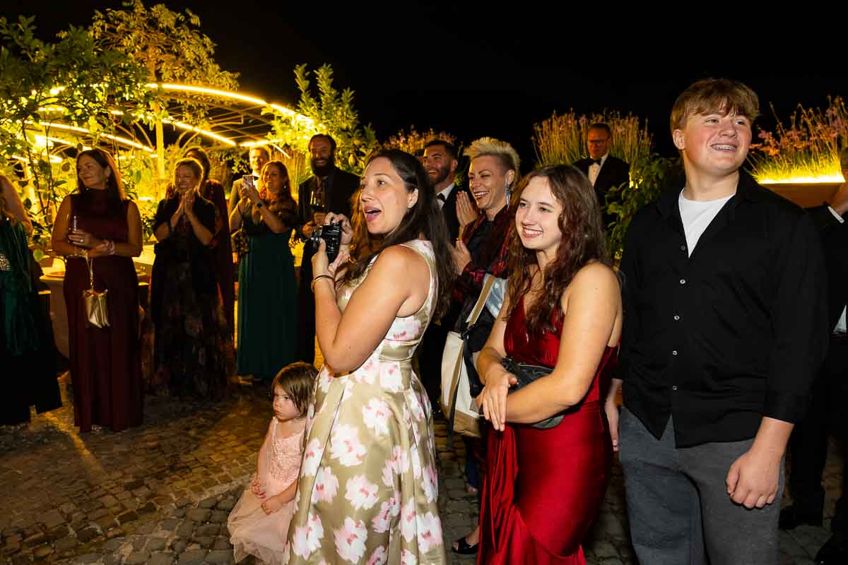 Excited friends and family members using their phones to capture the cake cutting moment.